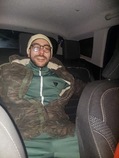 A single adult male is depicted seated in the rear passenger compartment of a vehicle in Meknes, Morocco. The subject, wearing a light-colored beanie and round-framed eyeglasses, is looking directly at the camera with a broad smile, revealing his teeth. He is dressed in a dark green long-sleeved tracksuit top with a white zipper and a dark emblem on the left chest, layered under an unzipped, camouflage-patterned jacket with a light-colored fleece lining visible at the collar. His lower body is clad in dark green trousers, matching the tracksuit top. The vehicle's interior is dimly lit, featuring a light grey ceiling and dark fabric seats with a textured pattern visible on the front passenger seat. Two dark headrests are visible behind the subject. No other individuals or external features are discernible.