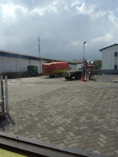 The image depicts an outdoor scene, likely a logistics or industrial area, under a cloudy sky. In the foreground, a paved area with interlocking rectangular bricks is visible, with some patches of water or dampness. To the left, a metal railing or fence post suggests the edge of a viewing area or walkway.
Further into the scene, two trucks are present. One is a green semi-trailer truck, seemingly empty and parked in the lot. The other truck, positioned to the right, is a flatbed truck with a yellow chassis and a cargo covered by a large, bright orange tarp. The truck with the tarp appears to be longer and more heavily loaded.
In the background, there are several structures. A long, white building with a blue trim runs along the left side of the image. To the right, a portion of a white building with a dark roof and a small window is visible. Between the buildings, there are glimpses of trees and other industrial-looking structures, including a tall telecommunications tower. A traffic light pole with red and white markings and a bright orange traffic cone in front of it is also situated near the trucks.
The overall impression is one of a functional area, possibly a loading dock or storage yard, with vehicles and infrastructure indicative of transportation and industry. Given the context of Surabaya, Indonesia, this scene is consistent with the industrial or port areas found in such a city.