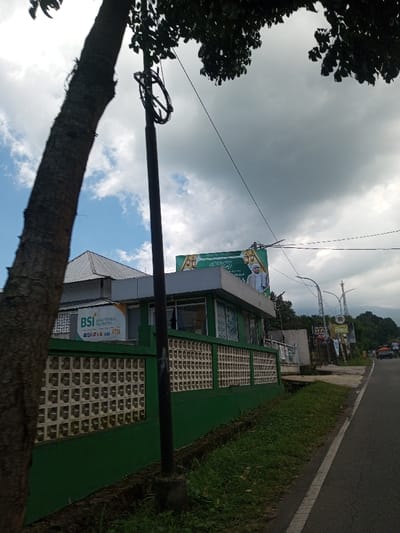 The image depicts an urban roadside environment, stated to be located in Tagtse Tashidingkha, Bhutan. In the foreground, a tree trunk stands on the left, with dark green foliage visible at the top. A dark utility pole, centrally positioned, features a coiled black cable near its apex. To the right of the pole, a building with a light grey roof and green exterior walls is visible. This building prominently displays a 'BSI BANK SYARIAH INDONESIA' sign, accompanied by VISA, MASTERCARD, GPN, and ATM logos. Below the building's windows, a green-painted concrete fence with square lattice cutouts extends along its perimeter. Above the building, a large green billboard features white text, including Arabic script and what appears to be Indonesian, alongside an illustration of a man wearing white traditional clothing and a white head covering. A paved road with a white edge line parallels the building on the right, extending into the distance where smaller signs, streetlights, and several vehicles are discernible. The background consists of distant green hills or mountains under a sky exhibiting both patches of blue and significant grey cloud cover. No live individuals are observed within the scene; a stylized male figure is depicted on the billboard.