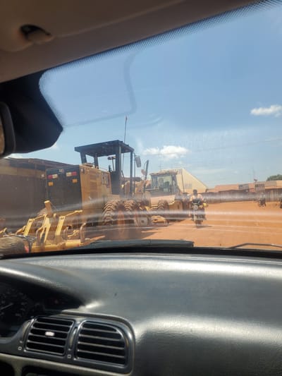 The image presents a view from inside a vehicle in Yelwa, Nigeria, looking out through a windshield with visible smudges and watermarks. The vehicle's dark gray dashboard occupies the foreground, displaying air vents on the left and a partial view of the instrument cluster. The upper edge of the windshield shows a dotted frit band, and a section of the vehicle's light-colored headliner is visible at the top.
The outdoor scene is dominated by heavy construction equipment on a reddish-brown dirt surface, indicative of an unpaved road or construction site, under a clear blue sky with scattered white clouds. A prominent yellow Caterpillar (CAT) motor grader is positioned in the mid-ground, observed from its rear-left, with its large tires, engine compartment, and open operator's structure clearly visible. The 'CAT' logo is discernible on its side. To the right and slightly behind this grader, another yellow piece of heavy machinery, possibly a second grader, is partially visible.
Several motorcyclists are present on the dirt road. One individual on a motorcycle, wearing a yellow or orange vest, is centrally located to the right of the main grader, moving away from the viewer. Other motorcyclists are visible further in the background to the right. In the distant background, power lines traverse the frame, and a large, light-colored building with a sloped roof stands behind the machinery, alongside other smaller, indistinct structures on the horizon.