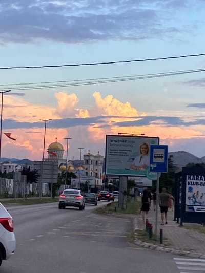 This is a photograph taken in Bar, Montenegro, likely during the late afternoon or early evening, judging by the soft, warm tones of the sky and the low angle of the sunlight illuminating the clouds. The scene depicts a street with a sidewalk and several cars.
The foreground features a portion of a white car, possibly at a bus stop, with its taillights illuminated. Further down the road, more cars are visible, including a white SUV with its brake lights on. The road itself has dashed white lines marking the lanes, and a pedestrian crossing is visible on the right side of the image.
Along the sidewalk, there are several streetlights and billboards. One billboard advertises "KARAT KLUB-BA," suggesting a martial arts club. Another prominent billboard displays a bus stop sign above an advertisement featuring a smiling woman and text in what appears to be Cyrillic and Latin script. Part of this text is legible as "Stručnost boja" and "Cirineco/vanna."
In the background, several buildings with distinctive architectural features, including domes and minarets, are visible. One building has a golden dome topped with a cross, hinting at a religious institution. The overall impression is of a town or city transitioning into evening, with a mix of everyday urban life and cultural elements. The sky is filled with dramatic clouds, colored by the sunset, creating a serene and visually striking atmosphere.