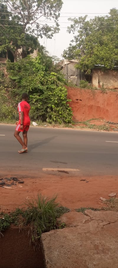 The image presents an outdoor scene in Ogidi, Nigeria, dominated by a paved road running horizontally across the mid-ground. A young individual, appearing male, walks from left to right across the dark grey asphalt road, facing slightly away from the camera. He wears a red short-sleeved athletic shirt with white stripes on the sleeves and "JAJA ABUBAKAR HOUSE" printed in white on the back, along with matching red shorts featuring white side stripes. Light-colored flip-flops are on his feet, and his shadow extends to his right on the road surface.
The road exhibits faint white and yellow markings and some dark stains. On both sides of the road, the ground consists of reddish-brown earth. The immediate foreground on the left features sparse green weeds and scattered dark debris. On the right foreground, a broken concrete slab borders a visible drainage channel, surrounded by green grass and weeds.
Beyond the road, the background features a steep reddish-brown dirt embankment on the right, topped by lush green foliage, several trees, and rudimentary structures, including a grey water tank and buildings with corrugated metal or tarpaulin components. To the left and behind the individual, dense green trees and bushes partially obscure what appears to be a multi-story building. The sky overhead is light grey and hazy.