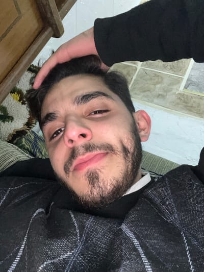 A man with dark hair, eyebrows, and a beard looks at the camera. He wears a black turtleneck and a plaid gray jacket. A hand rests on the back of his head, seemingly holding his hair. The background consists of a textured white wall, a carpet with yellow and brown patterns, and a wooden chair. The photo is taken at an angle, tilting the subject's head.