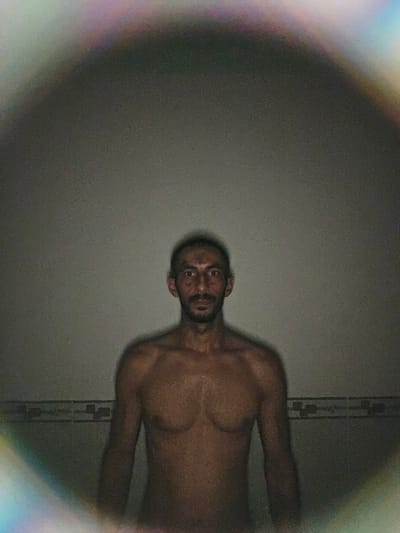 The image displays a shirtless man, positioned horizontally within the frame, looking directly at the camera. His body is oriented with his head towards the upper right and his torso extending towards the lower left. He has dark skin, a beard, and a neutral to slightly surprised expression, characterized by wide eyes and a slightly open mouth.
The background consists primarily of a plain, dark gray wall. On the left side, a narrow section of a tiled wall is visible, showing a subtle pattern. The lighting is dim, resulting in a noticeable grainy texture across the entire image. A prominent lens flare effect is present around the perimeter of the frame, creating a colorful, rainbow-like glow with discernible hues of purple, blue, green, and red, particularly along the top and right edges. This effect suggests a direct light source interacting with the camera lens. No text is visible in the image. The indoor setting and artificial lighting prevent determination of the time of day.