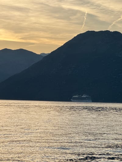 The image depicts a wide shot of the Bay of Kotor near Dobrota, Montenegro, at sunset. The sky is filled with wispy clouds illuminated by the warm hues of the setting sun, ranging from pale yellow to a soft orange. In the foreground, the calm sea reflects the golden light, creating a textured surface of ripples. Dominating the mid-ground is a large white cruise ship, positioned to the right of the frame. Behind the ship, a series of dark, silhouetted mountains rise steeply, their rugged forms contrasting sharply with the luminous sky. The overall scene is serene and picturesque, capturing the natural beauty of the Montenegrin coast during twilight.