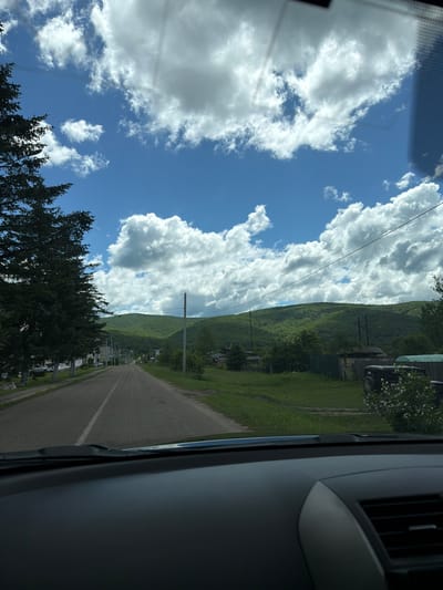 The image shows a view from inside a vehicle driving on a paved road. The road leads into a village nestled between rolling green hills. The sky is a vibrant blue, dotted with fluffy white clouds. On the left side of the road, there are houses and a tall evergreen tree. On the right side, there are more houses, fences, and vegetation. In the distance, the hills are covered in dense green trees. The scene appears to be during the daytime, with good visibility. The location is identified as the city of Kul'dur, Russia.