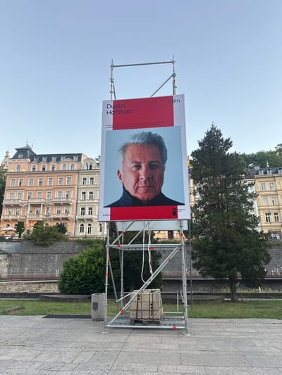 An outdoor publicity display for the 60th Karlovy Vary International Film Festival features a large portrait of actor Dustin Hoffman. The display is supported by scaffolding and positioned on a paved area adjacent to a grassy embankment. In the background, several historic multi-story buildings with ornate facades are visible. A dense evergreen tree stands to the right of the display, partially obscuring the buildings. The sky is clear and blue, indicating daytime. A trash receptacle and a vintage suitcase are placed near the base of the scaffolding.