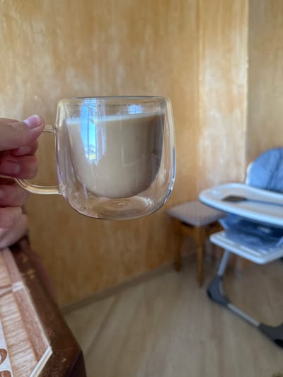 The media file shows a close-up, side-view of a clear, double-walled glass mug filled with a light-colored beverage, likely coffee or tea, being held by a person's hand. The mug has a transparent handle. The background is out of focus, but appears to be a warm-toned, wooden surface, possibly stairs or a shelf. In the upper portion of the background, there are hints of household items, including a blue fabric and white plastic structures, suggesting an indoor domestic setting. The lighting suggests it could be daytime, with a natural light source reflecting off the mug. There are no visible text elements in the image. The context provided indicates the location is Chemerne, Ukraine. The image is candid and focuses on the beverage and the hand holding the mug.