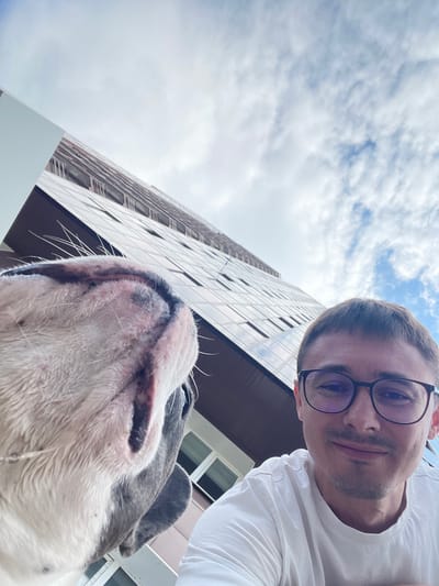 In a low-angle, wide-shot selfie, a man with short brown hair, glasses, and a light-colored t-shirt smiles at the camera. He is partially obscured by a dog's head in the foreground, which is positioned very close to the lens, with its snout, mouth, and whiskers prominently displayed. The dog appears to be white and grey. Behind the man and dog, a tall, modern building with a glass facade rises steeply towards a cloudy sky. The architecture of the building, characterized by its reflective surfaces and repeating window patterns, suggests an urban setting. The overall scene captures a moment of playful interaction between the man and his dog, with the imposing city architecture serving as a backdrop. The image was captured in Novosibirsk, Russia.