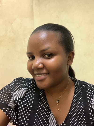 A woman with dark skin and black hair is smiling at the camera. She is wearing a black and white polka dot dress and a gold necklace with a butterfly pendant. The background is a yellowish-tan wall. There is no information about the location being in Dar es-Salaam, Tanzania.