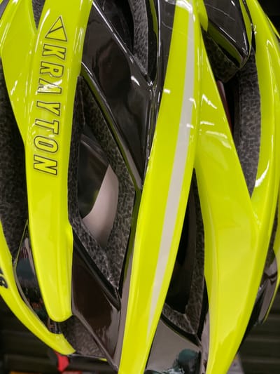 The image shows a close-up of a bright yellow bicycle helmet. The helmet has a glossy finish and black accents, with a prominent white stripe on one side. The brand name "KRAYTON" is visible in raised, silver lettering on the left side of the helmet, with a small triangle above the "K." The helmet's interior padding is visible in some of the vents, appearing to be grey and textured. The lighting suggests an indoor setting, possibly a retail display or a storage area. The image does not provide any information or visual cues to determine a specific location in the city of Málaga, Spain.