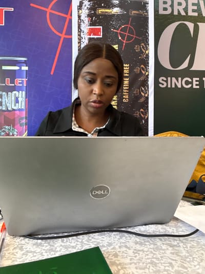 A photograph taken indoors in what is likely Itirin, Nigeria, contains a woman, packaging materials, and a Dell laptop. The woman is turned to her right, positioned between the product packaging and the laptop, looking at the camera. The packaging appears to be for beverage products, with text fragments like "BREW CO" and "CAFFEINE FREE" visible. The Dell laptop is open, angled towards the top-right, and the Dell logo is prominently visible. A cord is plugged into the laptop, and a patterned surface is beneath it. Part of a green object is visible to the bottom-right of the laptop.