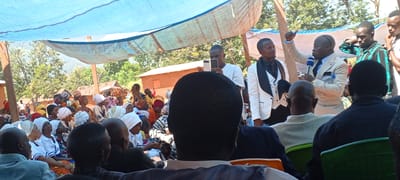 The scene depicts an outdoor gathering under a makeshift canopy, likely in a rural setting near Kalemie, Democratic Republic of the Congo. A group of people, predominantly women wearing colorful headscarves, are seated and facing a group of men who appear to be speaking. One man in a light-colored suit is holding a microphone and gesturing with his hand. Another man in a white suit is standing next to him. A third man is holding a smartphone, seemingly recording the event. The audience is composed of people of varying ages. The environment includes simple buildings with thatched roofs and lush vegetation in the background. The setting is bright, with natural light illuminating the scene.
