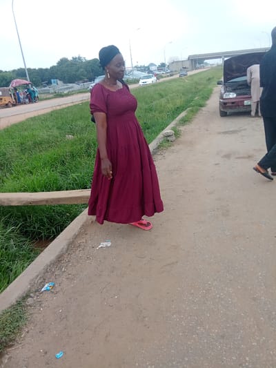 In Kagini, Nigeria, an outdoor scene unfolds by a dusty road lined with green grass. A woman, wearing a maroon dress and a black headscarf, stands on the edge of a concrete path. Her dress has a gathered bodice and a tiered skirt that reaches her feet, where she wears pink flip-flops. A wooden plank is visible in the foreground, running horizontally. To the right, a maroon car with its hood open is being examined by two men. One man, wearing a pale garment, is bent over the engine. The other man, wearing dark clothing, stands beside him. In the background, a highway overpass can be seen, with vehicles moving on it. Further in the distance, there are buildings and trees. A yellow three-wheeled vehicle, possibly a tricycle taxi, is parked near some stalls with awnings. The sky is overcast. The ground is littered with some trash.