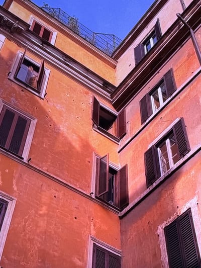 The image shows an upward angled view of the exterior of buildings. The buildings are orange and peach colored, with visible signs of wear and tear on the stucco walls. Several windows with dark brown shutters are visible, some open and some closed. A metal railing and some greenery are present at the top of one of the buildings, suggesting a rooftop area. The sky is a clear, bright blue. The image does not provide any specific details to indicate that the location is Dubai, United Arab Emirates.