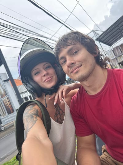 The image shows two individuals, a man and a woman, in Munggu, Indonesia, both appearing to be of Caucasian descent. The woman is wearing a black motorcycle helmet with a clear visor, and a light-colored top. Her chest features a floral tattoo. A large tattoo is visible on her arm as well. The man is wearing a red t-shirt. They are close to each other, likely on a motorcycle. Background elements include overhead power lines, buildings, and potentially a vehicle.