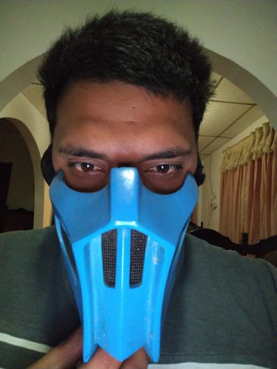 A close-up, eye-level portrait shows an individual, likely an adult male, wearing a bright blue mask over their nose and mouth. The person has dark, short hair and visible brown eyes that look directly at the viewer. Their skin tone is medium. The mask is made of plastic, featuring distinctive angular shapes and black mesh inserts where the mouth would be. It appears to be held in place by ear straps, with a hand or thumb visible at the bottom of the mask.
The individual is wearing a dark green t-shirt, with a light-colored stripe (possibly white) visible towards the bottom left. The background suggests an indoor setting, likely a home in Tinaquillo, Venezuela. Two arched openings are visible: one on the left behind the person's head, and another on the right. Through the right arch, light brown or beige curtains are visible, suggesting a window or another room beyond. The lighting is soft and ambient, consistent with an indoor environment. No text is visible in the image. The photo captures a casual pose with the distinctive mask.