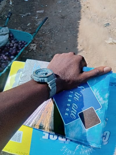 The image shows a close-up view of a person's dark-skinned arm and hand holding a stack of notebooks. The arm is positioned diagonally across the frame, extending from the lower left towards the upper right. A light-colored, possibly grey or light blue, digital wrist-watch is visible on the person's wrist. The hand is holding several notebooks, with the thumb resting on the top of the stack.
The most prominent notebook is blue with white text that appears to read "8 in one NOTEBOOK". It features two overlapping square shapes, one blue and one dark brown or black. Beneath this, other notebooks are visible. One has "JOKEL-EAGLE" printed along its side, and another yellow-colored notebook has text "Agboyim" and what looks like a barcode on its visible edge.
The background consists of a dusty, light brown or beige ground surface, suggesting an outdoor setting. A distinct shadow from the arm and notebooks is cast on the ground. In the lower-left corner of the image, part of a light blue or green wheelbarrow is visible, containing some dark, possibly patterned fabric or clothing. The lighting is bright, indicating it is daytime, likely under direct sunlight, which creates strong highlights on the notebooks and sharp shadows on the ground.