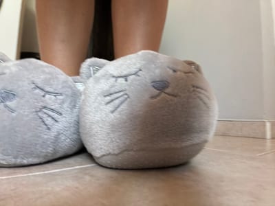 A close-up, low-angle shot shows a pair of fuzzy, gray cat-shaped slippers resting on a tiled floor. The slippers have embroidered closed eyes and whiskers, and the one on the right has a small, dark gray nose. The feet and lower legs of a person, wearing dark clothing above the knees, are visible in the background, behind the slippers. The floor is made of light-colored square tiles with thin white grout lines. A white baseboard runs along the wall in the background. The scene is indoors, and the lighting appears to be natural, coming from the right side of the frame. There is no information to suggest this image is located in Novi Iskar, Bulgaria.