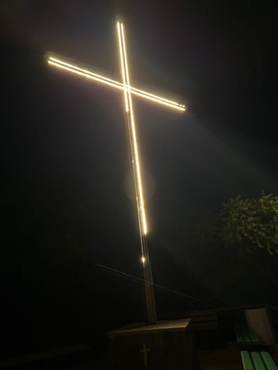 A large, illuminated cross stands tall against a dark, night sky. The cross is outlined with bright, golden-yellow LED lights, which create a striking contrast with the surrounding darkness. The vertical beam of the cross extends upwards, while a horizontal beam extends to the left. A faint mist or fog appears to be present in the air, catching the light and creating a soft glow around the cross. To the right and slightly behind the cross, the dark silhouettes of trees are visible, with some foliage illuminated by the cross's light. In the foreground, at the base of the cross, a concrete structure is present, also dimly lit. The overall scene suggests a nighttime illumination of a religious symbol, possibly in a park or outdoor religious site. This illuminated cross is a prominent landmark in the city of Asenovgrad, Bulgaria, located on a hill overlooking the city.