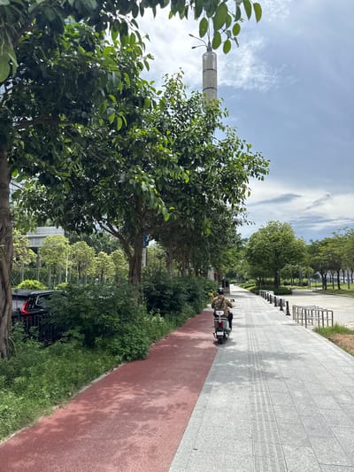 In Haizhu District, China, a person rides a scooter on a paved pedestrian walkway adjacent to a red-colored bicycle lane. The walkway is lined with trees and decorative bollards. In the background, a tall, cylindrical tower with a patterned surface stands against a partly cloudy sky. The scene depicts a well-maintained urban environment with a focus on pedestrian and cyclist infrastructure.