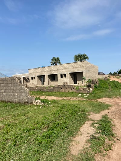 The image depicts an unfinished, multi-story building constructed from concrete blocks in Kuson, Nigeria. The building has numerous open window frames and doorways, suggesting it is in the early stages of construction. Two palm trees are visible, one taller than the other, with the taller one appearing to be near the side of the building. The sky above is a clear blue with some wispy clouds. To the right of the building is a patch of green grass and a dirt path. The scene appears to be an outdoor setting under a bright sky, likely during the daytime. There are no people visible in the image. The overall impression is one of ongoing construction in a rural or semi-rural environment.