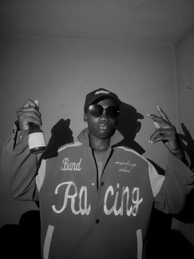 This is a black and white, vertically oriented portrait of a young person, likely male, taken indoors in Asaba, Nigeria. The subject, with dark skin, wears sunglasses, a dark cap, and a two-toned jacket with contrasting panels and embroidered text. The jacket prominently features "Blind" (or "Bind") on the left chest area and large script spelling "Ra Cing" (suggesting "Racing") across the torso. Smaller text reading "motois club" (likely "motors club") is visible on the right chest.

The person is posing confidently, looking directly towards the camera, though their eyes are obscured by dark sunglasses. Their right hand is raised with fingers splayed, casting a strong, angular shadow on the wall behind them. In their left hand, they hold a dark bottle with a white label. Partially legible text on the label suggests it is a pharmaceutical syrup bottle, possibly containing "Codeine," and a QR code is also visible.

The setting is a simple, sparsely furnished room with plain, light-colored walls, forming a corner in the upper left background. Strong, directional lighting creates prominent and sharp shadows of the subject's head and raised hand against the wall, adding a dramatic effect to the monochromatic scene. The overall impression is one of street style or a posed fashion shot, with the subject's attire and accessories being key elements.