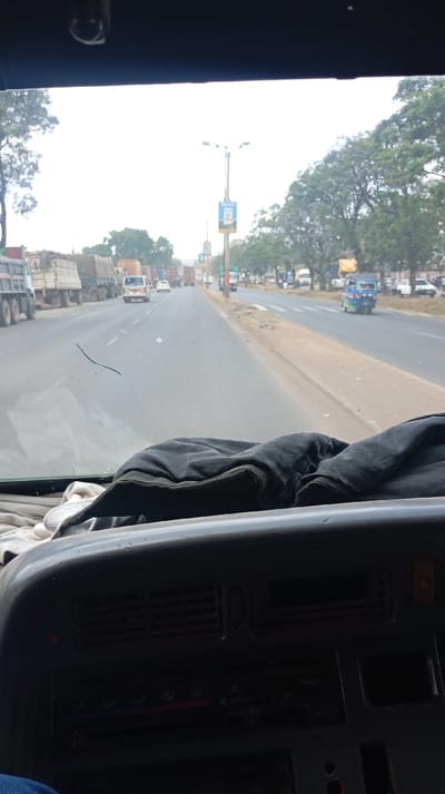 A view from inside a vehicle shows a straight road with multiple vehicles in the distance. Large trucks line the left side of the road. Trees are visible on both sides, with light poles along the median. The dashboard of the vehicle occupies the foreground, partially covered with a black garment. The weather appears overcast or hazy.