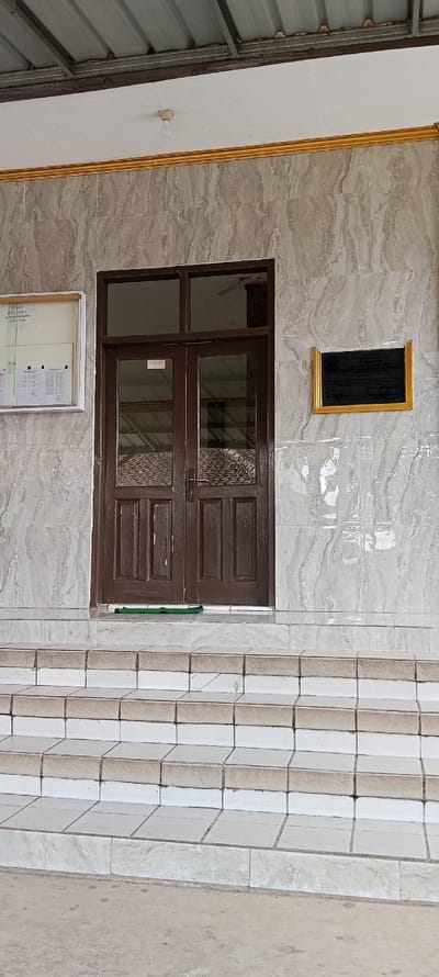 The image shows the front entrance to a building in Kecamatan Kutawaringin, Indonesia. The building is made of white marble tiles with a gold trim. The entrance has a set of double wooden doors with glass panels, and a small green mat in front of them. The doors lead to a hallway. There are five steps leading up to the door. There is a light fixture on the ceiling above the entrance and a small plaque on the wall to the right of the doors. The plaque is gold-framed and contains a black background. There is a bulletin board to the left of the doors with a white background and a black frame. The surrounding environment is a concrete walkway, a light grey in color.  There are no people or objects in front of the door.