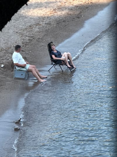 This media file depicts two individuals at the edge of the water in Boreti, Montenegro. The scene is a beach with a sandy shore meeting the gentle waves of the sea. The water appears clear and shallow near the shore.

The first subject, a woman, is seated in a portable chair, facing towards the viewer and smiling. She is wearing a dark blue patterned dress. Her chair is positioned on the wet sand, close to the water's edge.

The second subject, a man, is also seated in a similar portable chair. He is wearing a white t-shirt and light-colored shorts, and his feet are in the shallow water. He is facing away from the viewer, looking out towards the sea or the camera is angled to catch the back of his head. His chair is also on the wet sand.

The overall activity appears to be relaxation or enjoying the beachside. The lighting suggests it might be late afternoon or early evening, given the soft light and the warmth of the sand tones. The weather seems pleasant, with calm water and no obvious signs of wind or rain. The presence of portable chairs indicates a leisurely outing. The scene is set in a coastal environment, consistent with the location context of Boreti, Montenegro, known for its beaches. There is no visible text in the image.