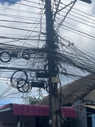 This is a medium-angle, eye-level, vertical shot of a utility pole heavily laden with a chaotic tangle of electrical wires and cables. The pole, made of concrete, is centrally located and extends out of frame at the top and bottom. Several large, coiled wires are wrapped around the pole, some forming perfect circles and others appearing haphazardly looped. Smaller, thinner wires extend from the pole in every direction, creating a dense network against a cloudy, bright sky.
Beneath the pole and to the right, the roofline of a building is visible, covered with grey tiles. A portion of a pink awning or canopy stretches across the lower part of the image, beneath the chaotic wires. Text is partially visible on the awning, with "EYELAS" and "NAILS" discernible. To the right of the awning and building, a corrugated metal structure is visible.
The scene appears to be in Kamala, Thailand, given the context of previous images in this series, suggesting an urban or semi-urban environment with overhead power lines. No people are visible in the image. The focus is on the dense, jumbled network of wires, highlighting a common characteristic of utility pole infrastructure in some areas. The lighting is bright but overcast, indicating daylight conditions.