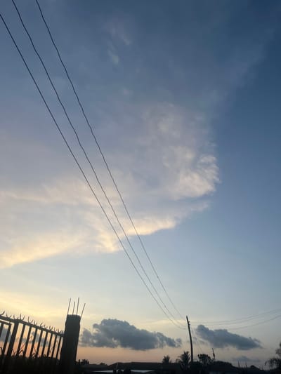 This image captures an outdoor scene in Barakin Akawo, Nigeria, during either sunrise or sunset, indicated by the warm, transitioning light in the sky. The sky occupies the majority of the frame, displaying gradients from pale orange-yellow and light blue on the left to a deeper azure on the right. Varied cloud formations are present: ethereal, wispy clouds with subtle warm coloration are distributed across the mid-left and center of the sky, while a more concentrated, darker grey cloud mass is visible above the horizon on the lower-right side.

Four dark, undulating power lines traverse the sky diagonally from the upper left towards the lower right. A silhouetted utility pole stands on the right side of the frame, with additional power lines extending from it.

The lower portion of the image reveals a silhouetted horizon line. In the foreground on the left, a section of a dark metal fence with pointed spikes is visible. Adjacent to the fence stands a concrete pillar with four exposed rebar rods projecting vertically from its top. Below the horizon, indistinct dark shapes represent buildings, other structures, and several trees, some appearing to be palm trees. No human figures or active interactions are discernible within the frame.