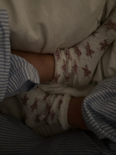 The scene is located in a dimly lit indoor environment, specifically depicting a bed in Wealden, United Kingdom. Two human lower legs/feet are visible, resting. Both feet are clad in fluffy white socks featuring a repeating pattern of reddish-brown/maroon bow-like or cross-like motifs. Portions of a blue and white striped fabric, consistent with sleepwear or bedding, are present on both the left and right sides of the frame, partially covering the legs and blending with the surrounding light-colored bedding material, likely a duvet or sheet. No faces or specific individuals are identifiable; the focus is on the feet and immediate bedding. The overall impression is one of repose.