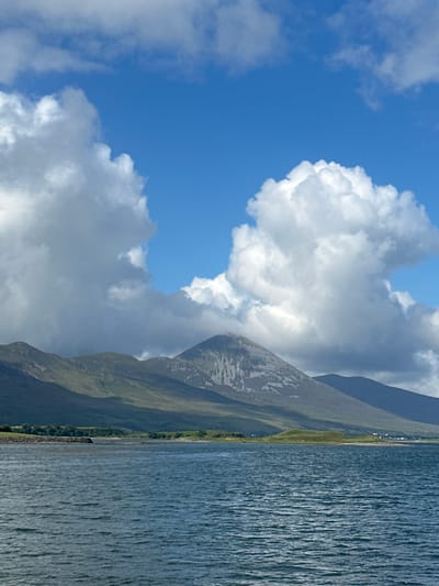 An expansive view of a mountain range with a prominent central peak rises behind a body of water, likely a bay or inlet, in Ireland. The sky is a vibrant blue, dotted with large, fluffy white clouds that cast shadows on the mountains. The mountains themselves are covered in green vegetation, with patches of bare rock visible on the slopes of the central peak. A thin line of trees and some scattered structures are visible along the shoreline. The water in the foreground is a deep blue-green, rippling with gentle waves. The overall scene evokes a sense of natural grandeur and tranquility.