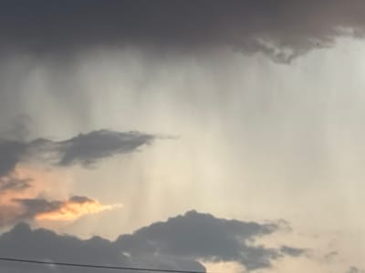 The image shows a cloudy sky in Pchelnik, Bulgaria. Above is a large, dark grey cloud mass, and below are other grey clouds with patches of sunlight peeking through. Rain or streaks of precipitation are visible from the upper cloud. A power line is visible.