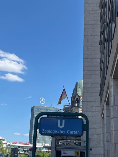 The image shows a bright, sunny day in Berlin, Germany, with a clear blue sky and a few wispy white clouds. The perspective is from below, looking upwards, and the image is rotated 90 degrees clockwise.
In the foreground, a blue sign with white text reads "U Zoologischer Garten". This indicates the Zoologischer Garten U-Bahn (subway) station, a well-known location in Berlin. The sign is mounted on a dark green metal frame.
Behind the sign, several modern glass and concrete buildings rise into the sky. One prominent building features a large white Mercedes-Benz logo on its facade. Another building has a distinctive architectural detail, possibly a historical tower or spire, with a green roof and intricate stonework. A German flag, with horizontal stripes of black, red, and gold, is visible on a flagpole.
The scene appears to be an urban environment, likely a busy street or plaza near the Zoologischer Garten station. The bright sunlight and clear sky suggest it's daytime, possibly late morning or early afternoon. There are no people clearly visible in the frame, but the presence of the U-Bahn sign and the buildings implies a public space. The overall impression is one of a vibrant, modern city under a pleasant sky.