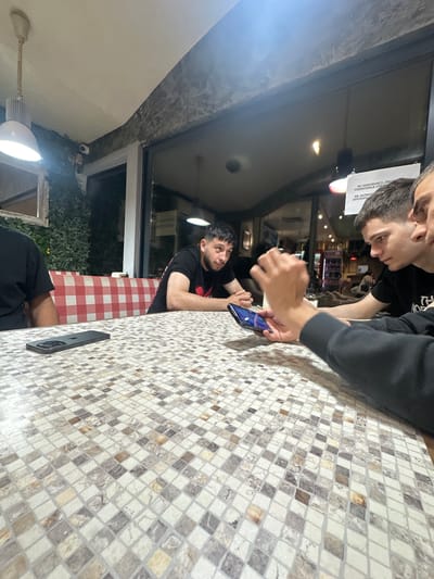 The image, taken in what appears to be an evening or nighttime setting, captures three men gathered around a mosaic-tiled table inside an establishment, possibly a cafe or restaurant in Silistra, Bulgaria.
In the foreground, an arm clad in a black long-sleeved top rests on the table, next to a black smartphone, likely an iPhone, lying face down. To the left, a man with dark hair and a beard, wearing a black t-shirt, is seated, looking upwards and to the side. Further back and to the right, another man, also in a black top, leans over the table, attentively viewing a smartphone he holds in his hands. His face is partially visible from above.
The table features a pattern of small, square mosaic tiles in shades of white, beige, and brown. In the background, beyond the seated men, a large glass window or door separates the interior from the outside darkness. Through the window, green foliage, possibly artificial, is visible, along with a small white security camera mounted high near the window frame. Another table in the background is covered with a red and white checkered tablecloth. Indoor artificial lighting illuminates the scene, with one distinct ceiling light fixture visible in the lower left of the frame.
A white paper sign is taped to the glass, displaying text in Romanian: "NU DESCHIDEȚI / ÎNCHIDEȚI", "FERESTRELE FARA", and "PROZOPTI". This translates to "Do not open / Close" and "Windows without / Blinds/Curtains" (or a similar meaning, "prozopți" being a less common word, possibly a typo for something like "jaluzele" or "perdele"). The overall atmosphere suggests a casual gathering.