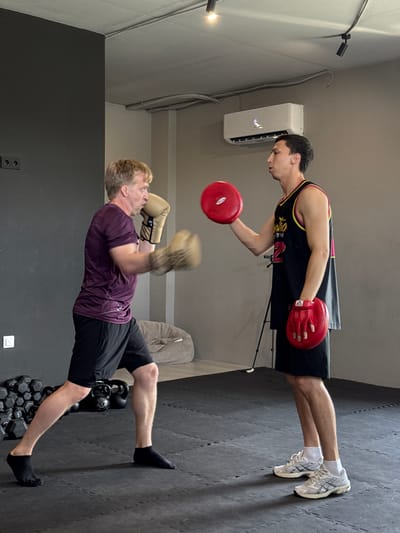 Two men are sparring in a gym. The man on the left, wearing a purple t-shirt and black shorts, is throwing a punch with boxing gloves. The man on the right, wearing a black and yellow tank top and black shorts, is holding up red boxing pads to block the punch. The gym has a dark gray wall on the left and a lighter gray wall in the background. There are weights and a kettlebell on the floor, and an air conditioning unit mounted on the wall. The floor is covered with black mats. This image is not in Tyumen, Russia.