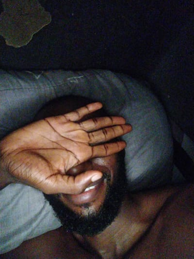 A close-up, dimly lit selfie shows a person's left hand covering their face. The hand is positioned over the eyes and forehead, with fingers spread. A portion of the person's mouth, showing teeth and lips, is visible below the hand, along with a dark beard. The person appears to be lying down, with a pillow visible beneath them. The background is dark and indistinct. The image appears to have been taken indoors, possibly in a bedroom, given the presence of a pillow and bedding. Without further context, it is impossible to determine the specific location within the city of Uyo, Nigeria.