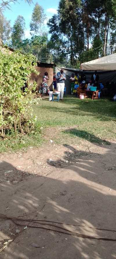 The scene is a gathering of people outside of a building in a rural area of Kipkarren, Kenya (latitude: 0.599, longitude: 34.957). A man is speaking into a microphone in front of a group of people seated on folding chairs in the shade of a white canopy tent. Another woman stands to the left in a long floral skirt, while other people sit on chairs to the right of the man. The man is wearing a long-sleeved gray shirt and gray pants, while the woman is wearing a long floral skirt and white shirt. The canopy tent is white with a brown border and is propped up on long poles. There is a table in the center of the scene with a white tablecloth and a blue bucket on it.  There are many trees surrounding the scene. There is a dirt path in the foreground. The ground is covered in green grass and some fallen leaves. The sky is blue with white clouds. The scene is in the middle of the day with a bright sky. The people are listening intently to the man speaking. There is an overall sense of peace and community.