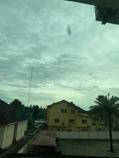 The image captures a partial cityscape in Ikeja, Nigeria, under an overcast sky with layered clouds. A tall, thin communication tower stretches upwards into the sky. A yellow building with multiple windows and air conditioning units is visible to the right, partially obscured by a palm tree. Below that, there's a building with a dark roof and a tan-colored exterior, and a pink outdoor umbrella. In the foreground, there are parts of buildings, and a patch of what looks like a parking lot. The image appears to be taken from an elevated perspective, likely from inside a building. A dark blob is visible in the sky, possibly a lens artifact.