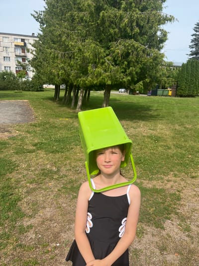 In Vangaži, Latvia, a young girl with a green bucket over her head stands in a grassy area. She is wearing a black swimsuit with white accents. Behind her, a row of coniferous trees lines a path, with a multi-story building visible in the background. The sky is overcast, and the ground is covered in dry grass. The scene appears to be a park or a residential area.