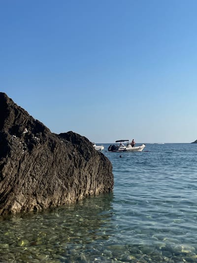 The image depicts a sunny day at a rocky coastline in Prijevor, Montenegro. The scene is divided by a large, dark, textured rock in the foreground on the left, and the clear, calm, blue water on the right. The water is shallow enough to see the pebble-filled seabed beneath, with sunlight creating dappled patterns on the rocks.

In the middle ground, a white motorboat is anchored in the water. Several people are visible on the boat, some appearing to be sitting or standing, engaged in various activities. One person is holding a phone. The presence of the boat and people suggests leisure activities like swimming or sunbathing.

The sky is a clear, bright blue, indicating fair weather and likely daytime, possibly late morning or early afternoon due to the strong sunlight. The overall atmosphere is peaceful and serene, characteristic of a summer day at the seaside. There is no visible text in the image.