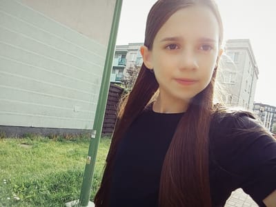 In the city of Dybów Kolonia, Poland, a young girl with long brown hair is captured in a selfie. She is wearing a dark t-shirt and her hair is styled with two low ponytails, with strands of hair catching the sunlight. Behind her, a pale green building with horizontal paneling is visible, and further in the background, modern apartment buildings with balconies rise against a bright sky. The ground is covered in green grass, and a tall green pole, possibly a street light or utility pole, stands to the left of the frame. The scene appears to be a bright, sunny day, possibly in a residential area.