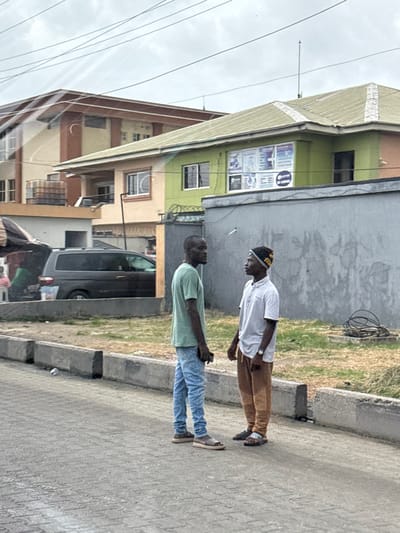 The image depicts a street scene in Alaguntan, Nigeria, under an overcast sky. In the foreground, two men of dark complexion stand on a paved road, facing each other, suggesting an interaction. The man on the left wears a light green t-shirt, light blue jeans, and brown sandals, holding a dark phone in his right hand. The man on the right wears a light-colored collared shirt, brown pants, dark sandals, and a patterned cap.
Behind the men, a low concrete curb separates the paved road from an unkempt area with dry grass and a coil of black cables on the right. To the left, a dark minivan is partially visible, parked behind a slightly elevated concrete section. Further left, a shaded stall with a partially visible individual is present.
In the background, two multi-story buildings are visible. The closest building has a light green upper floor with multiple informational banners displayed in its windows (e.g., "GLOBAL CERTIFICATES," "IT TRAININGS," "ACADEMY"), a lighter lower section, and a light-colored roof. This building is fronted by a grey concrete wall topped with barbed wire and a dark metal gate. To the left of this, a taller, multi-story building with a brown roof and light beige/cream walls with reddish-brown trim is visible, featuring balconies and a water tank on a lower roof section. Overhead, numerous dark power lines traverse the sky. A discarded silver can rests on the paved road near the curb.