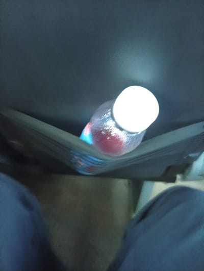 The image shows a close-up, overhead view of a clear plastic bottle with a white cap containing a red liquid, resting in a dark, fabric-lined compartment. The bottle is angled, with its cap pointing towards the top right of the frame. The fabric compartment appears to be part of a seat or bag. The background is a dark, textured surface. The lower portion of the image shows parts of blue fabric, likely clothing. The scene is indoors or within a vehicle. There is no indication of the specific location within Suk কুলovo, Russia.