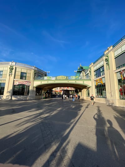 This is a photograph of the Prater amusement park in Vienna, Austria, taken on a sunny day. The image shows a street leading under an elevated walkway that is part of the park's architecture. The walkway is decorated with green trim and a sign that reads "PRATER". On either side of the street are buildings with shops and attractions. Several people are walking along the street and under the walkway. The sun casts long shadows across the pavement. The sky is clear and blue with a few wispy clouds. The overall scene depicts a vibrant and bustling atmosphere typical of a popular tourist destination.