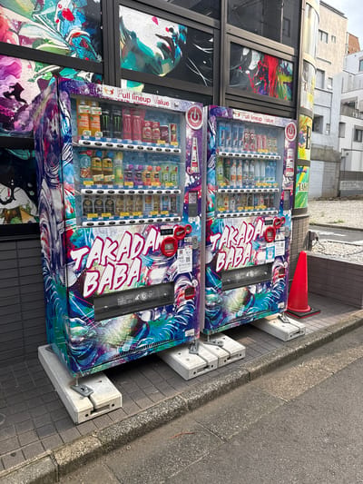 Two colorful, artistically wrapped vending machines are positioned side-by-side on a sidewalk in Shinjuku, Japan. The machines display a vibrant design featuring abstract art and text. The machine on the left has a wrap with a predominantly purple and blue color scheme, while the machine on the right has a similar wrap with variations in the design and color saturation. Both machines are elevated on concrete blocks for stability. The vending machines are filled with a variety of beverages, visible through the glass fronts. Price labels and product information are displayed on each item. The machine on the right has a red traffic cone placed next to it, on the edge of the sidewalk. The sidewalk is paved with gray tiles, and a paved street runs alongside it. In the background, there is a building with multiple windows, and another building with a light-colored facade. The overall scene depicts an urban streetscape with a focus on the uniquely decorated vending machines.