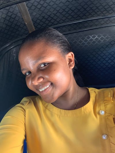 A young woman is posing for a photograph inside a vehicle. She is wearing a yellow blouse with pearlescent buttons at the collar and a delicate gold necklace. The woman has dark hair pulled back from her forehead, and she is smiling slightly, revealing her upper teeth. The background is a dark patterned surface, likely the interior of a seat or wall of the vehicle. The specific location in Dar es-Salaam, Tanzania cannot be determined from the image.