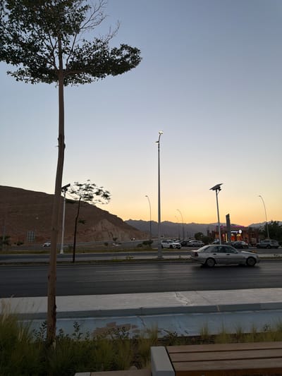 The image depicts an outdoor scene at dusk, likely in Sharm El Sheikh, Egypt, given the desert landscape and modern infrastructure. The sky is a gradient of pale blue transitioning to a warm orange and yellow hue near the horizon, suggesting the sun is setting.
The foreground features a large, leafless tree with a thick trunk on the left side of the frame. A portion of a wooden bench with a concrete base is visible on the far right, indicating a public space like a park or promenade.
The midground and background reveal a multi-lane road with several cars parked and driving. The road runs alongside a steep, rocky mountain range. Streetlights with solar panels are spaced along the road, illuminating the area. In the distance, several buildings with illuminated signs are visible, suggesting a commercial area. One sign appears to display colorful, possibly neon, lights, and another has text that might read "Sharm."
The overall impression is one of a developing urban area integrated with a natural desert environment, captured during a peaceful transition from day to night. The lighting suggests a clear, calm evening. There are no people visible in the image.